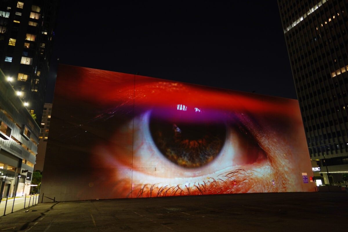 A monumental video projection onto a windowless wall of a five story building with downtown Los Angeles skyline in the background. The video shows an extreme closeup of a human eye filmed through a vibrant colorful array of screen reflections, it is ambiguous whether the eye is emanating light or receiving light.