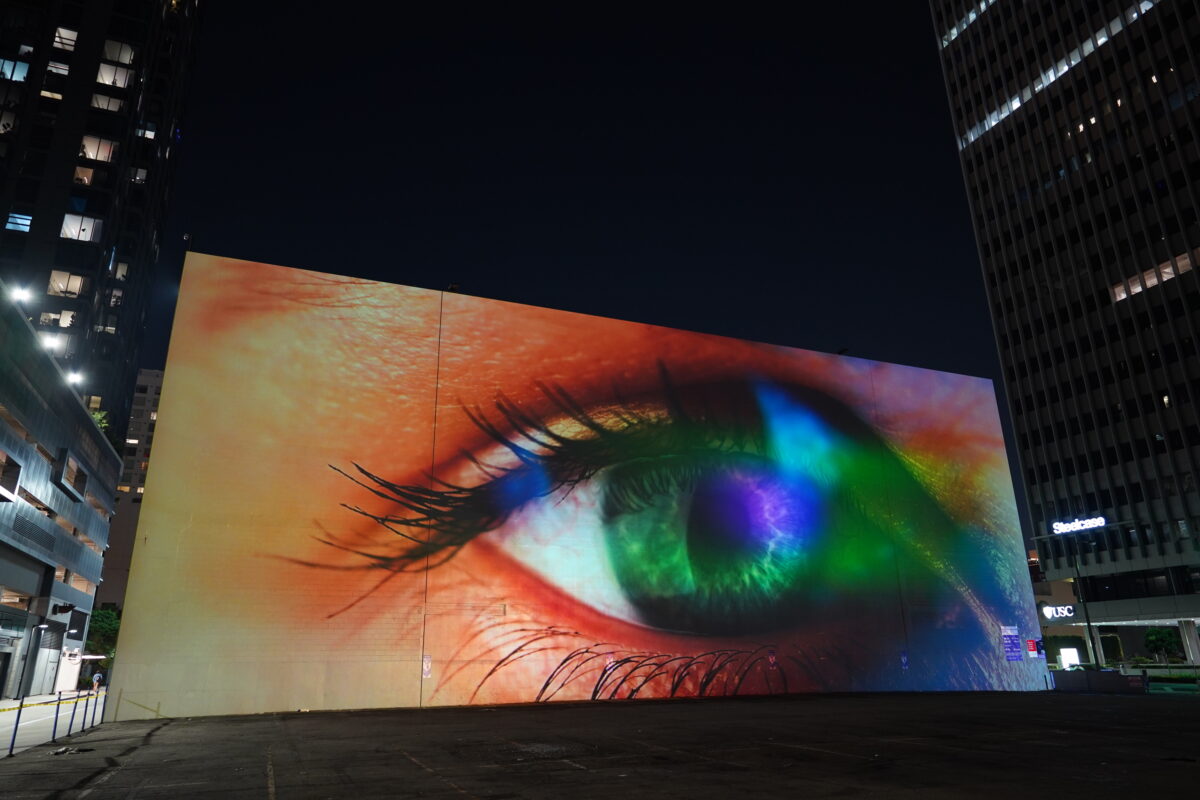 A monumental video projection onto a windowless wall of a five story building with downtown Los Angeles skyline in the background. The video shows an extreme closeup of a human eye filmed through a vibrant colorful array of screen reflections, it is ambiguous whether the eye is emanating light or receiving light.