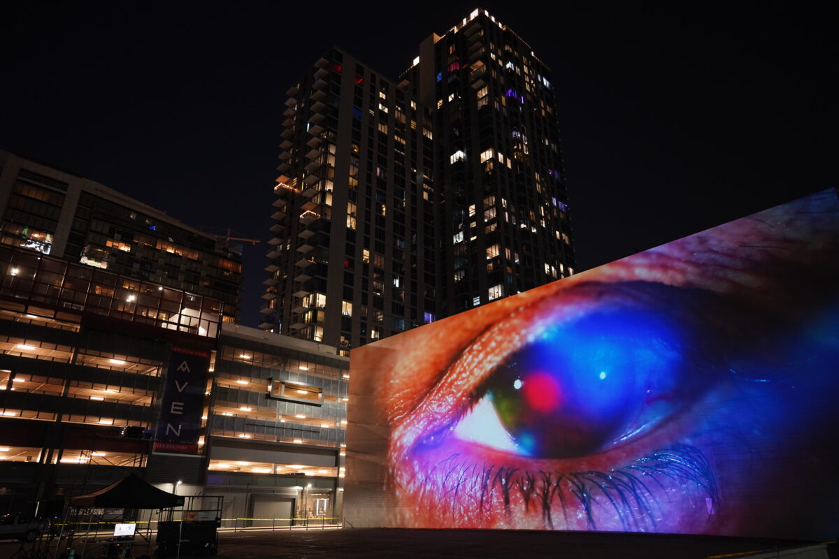 A monumental video projection onto a windowless wall of a five story building with downtown Los Angeles skyline in the background. The video shows an extreme closeup of a human eye filmed through a vibrant colorful array of screen reflections, it is ambiguous whether the eye is emanating light or receiving light.