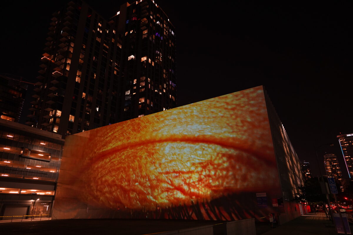 A monumental video projection onto a windowless wall of a five story building with downtown Los Angeles skyline in the background. The video shows an extreme closeup of a human eye filmed through a vibrant colorful array of screen reflections, it is ambiguous whether the eye is emanating light or receiving light.