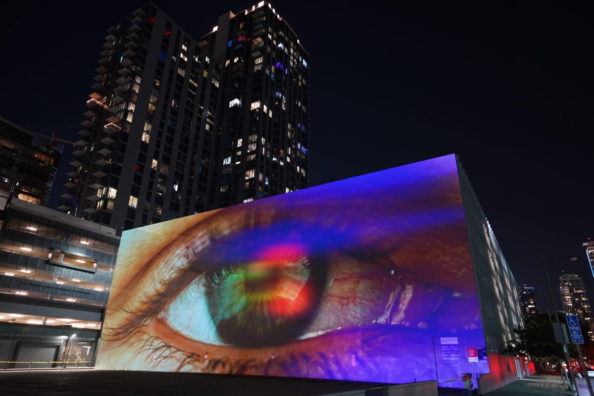 A monumental video projection onto a windowless wall of a five story building with downtown Los Angeles skyline in the background. The video shows an extreme closeup of a human eye filmed through a vibrant colorful array of screen reflections, it is ambiguous whether the eye is emanating light or receiving light.