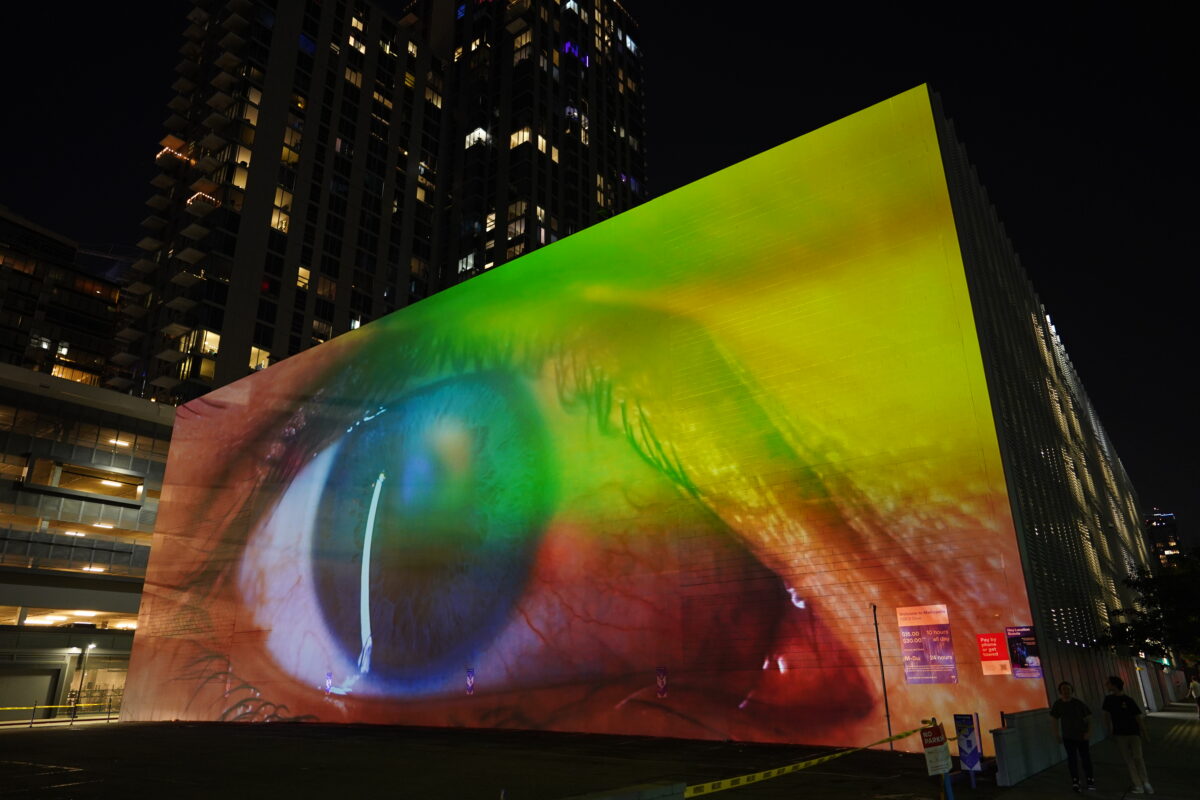 A monumental video projection onto a windowless wall of a five story building with downtown Los Angeles skyline in the background. The video shows an extreme closeup of a human eye filmed through a vibrant colorful array of screen reflections, it is ambiguous whether the eye is emanating light or receiving light.